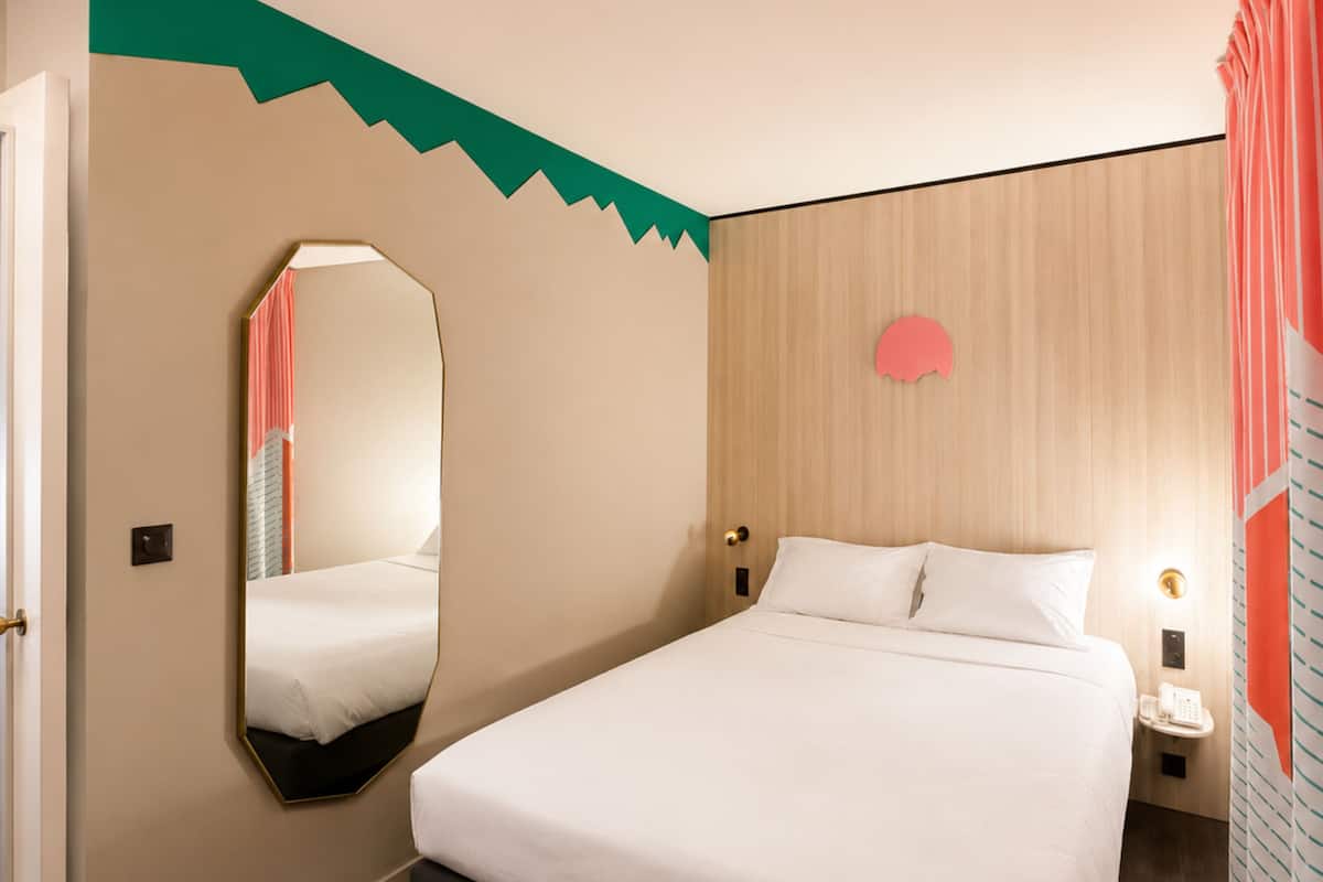 Una habitación de hotel con cama, espejo, mesita de noche con teléfono y lámpara de pared.