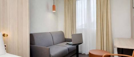 Et hotelværelse med en grå sofa, et lille bord med en bærbar computer, en træstol og en seng.