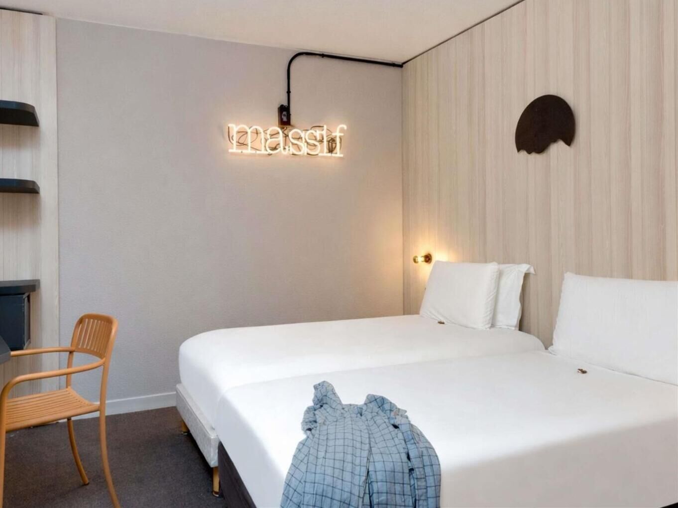 A hotel room with a bed, a chair, a wall-mounted light, and a decorative wall piece.
