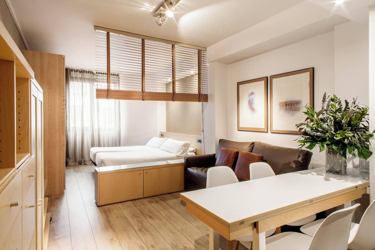 A compact hotel room with a bed, a sofa, a dining table, and a small kitchen area.