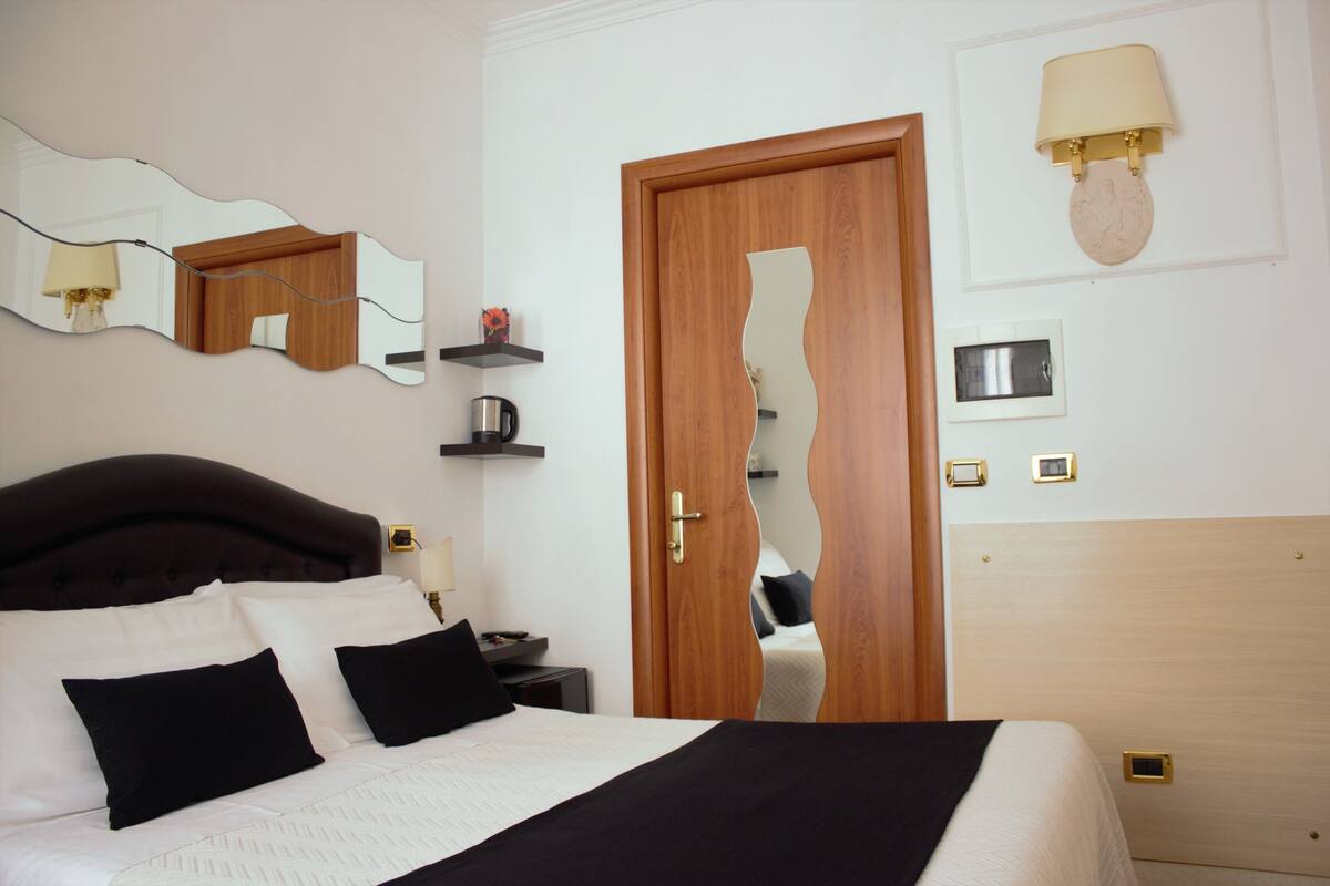 A hotel room with a large bed, a wooden door, a wall-mounted lamp, a shelf with a coffee maker, and a mirror.