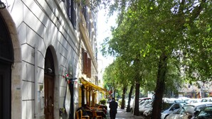 A street with parked cars, a sidewalk, and a building with a green awning and outdoor seating. - Città Eterna Bed & Breakfast (Rome)