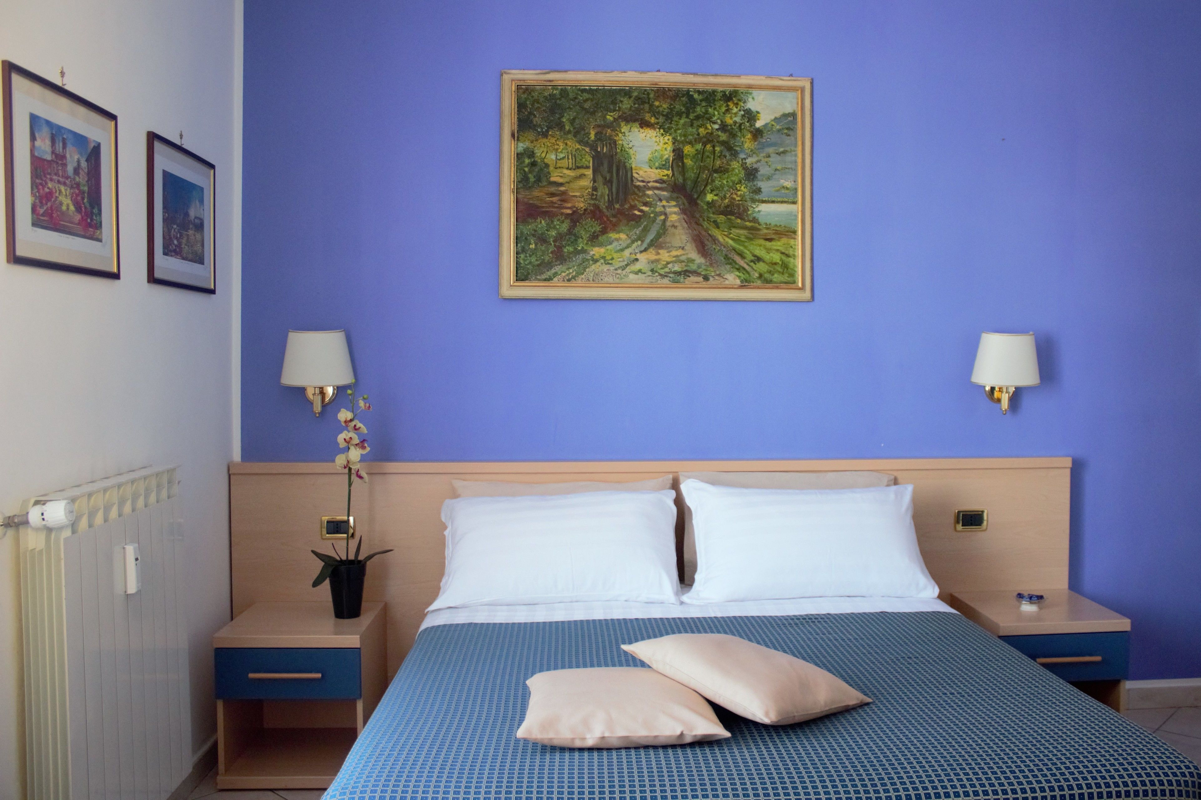 A hotel room with a bed, two bedside tables, a nightstand, a wall-mounted lamp, a framed painting, and a potted plant.