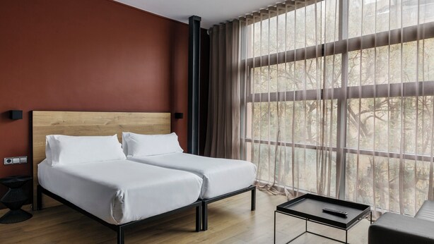 Ein modernes Hotelzimmer mit einem Bett mit Holz-Kopfstütze, einem kleinen Nachttisch, einer Couch und einem großen Fenster mit durchscheinenden Vorhängen.