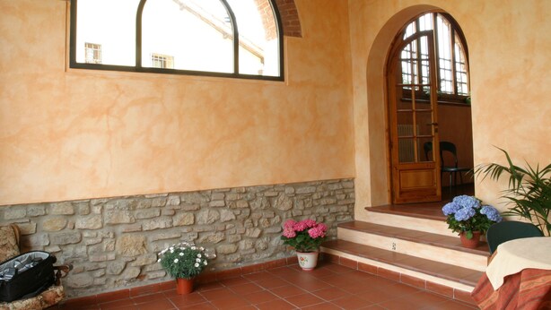 A room with a stone wall, a wooden door, and a window with a view of buildings. There are potted plants and a bench. - Il Colombaino (Scandicci)