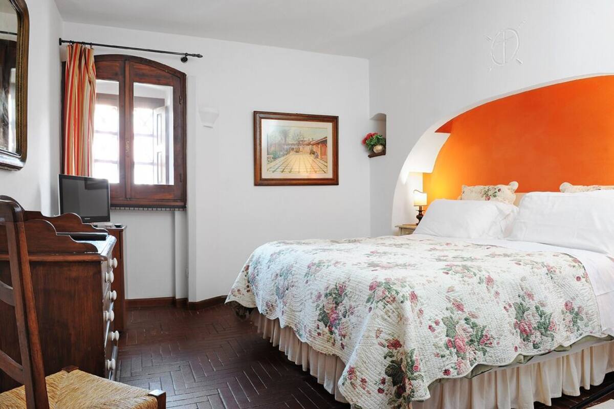 A bedroom with a wooden dresser, a bed with a floral bedspread, a framed picture on the wall, and a window with curtains.