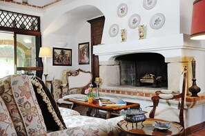 A traditional living room with a fireplace, a sofa, a table with books, and decorative plates on the wall. - Hotel Lucrezia (Riola Sardo)