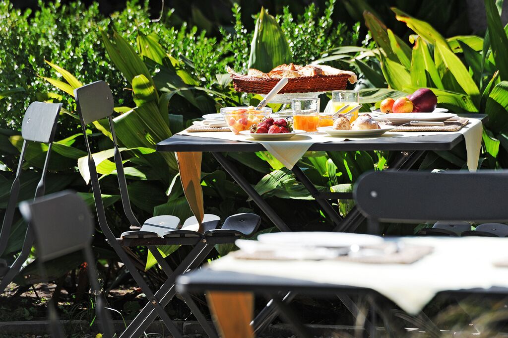 Un espacio exterior para comer con una mesa dispuesta para el desayuno, rodeada de sillas, en un exuberante jardín.