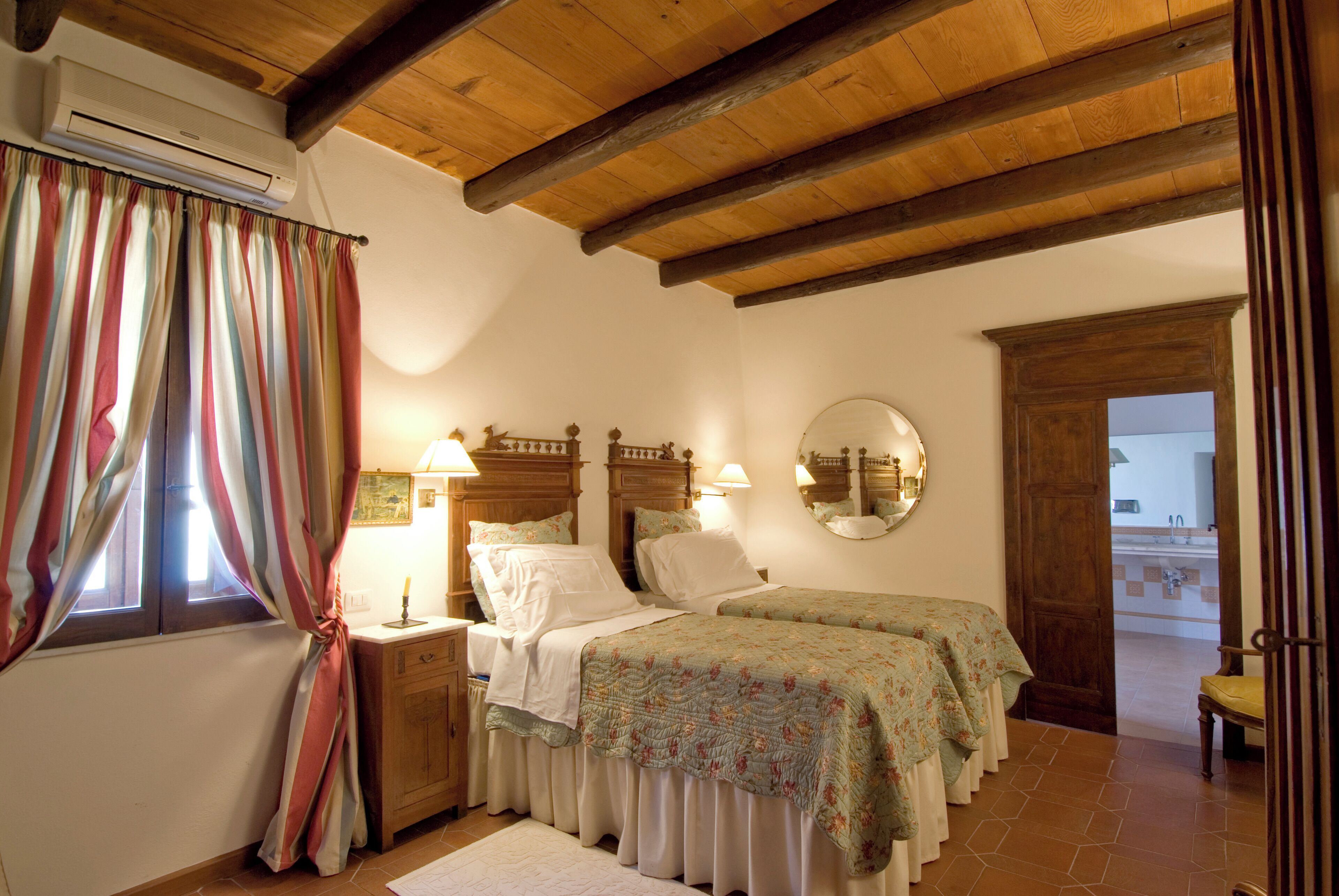 Un dormitorio con dos camas, vigas de madera en el techo, una ventana con cortinas, una mesita de noche con lámpara y un espejo en la pared.