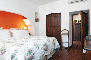 A bedroom with a floral bedspread, a wooden wardrobe, and a small shelf with plants. - Hotel Lucrezia (Riola Sardo)