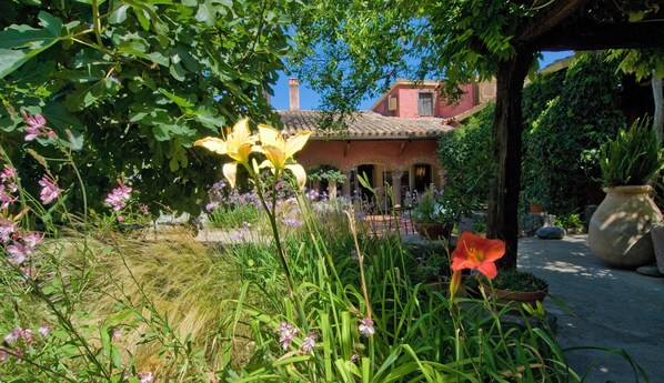 A garden with various flowers and plants, a building with a tiled roof, and a large pot on the ground. - Hotel Lucrezia (Riola Sardo)