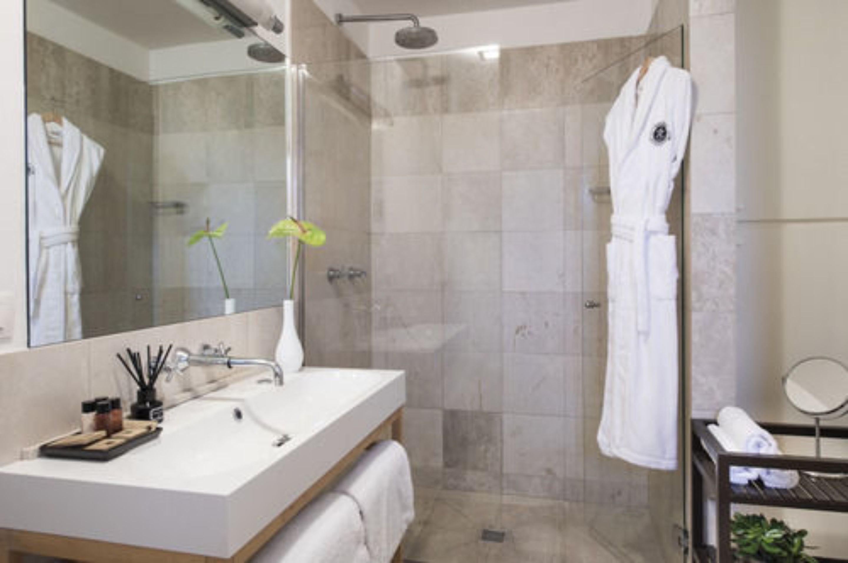 A modern bathroom with a large mirror, a sink, a shower, and two white bathrobes hanging on hooks.