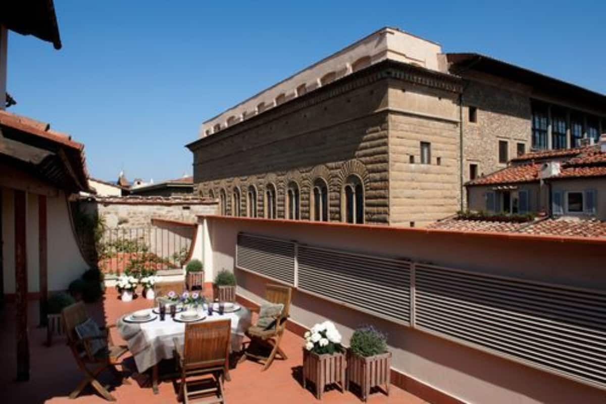 Terraço no telhado com uma mesa posta para refeições, cercado por vasos de plantas e uma parede de pedra.