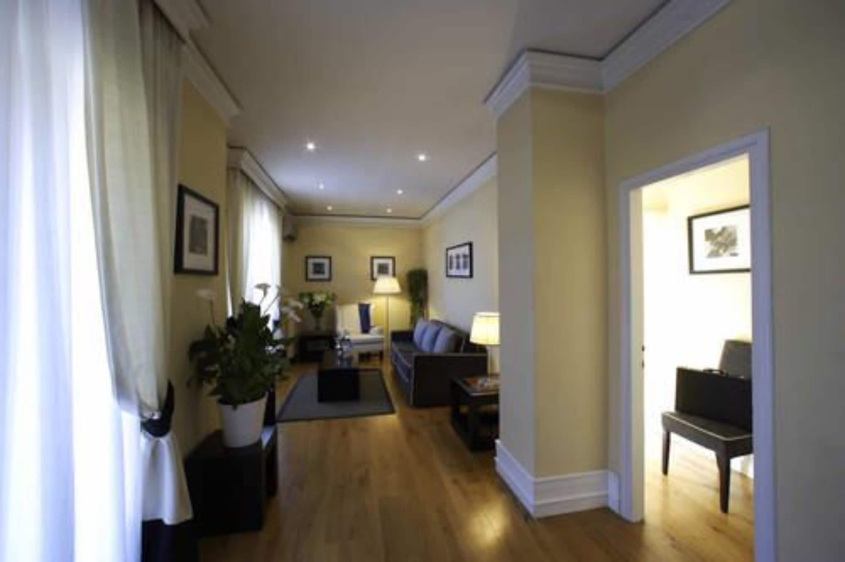 A well-lit hallway leading to a living room with a sofa, a coffee table, and a lamp. There are framed pictures on the walls and a potted plant on the left.