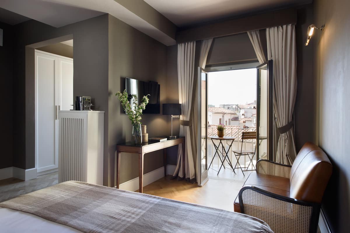 Quarto de hotel com cama, secretária com luminária, uma cadeira e vista da cidade através de amplas janelas.