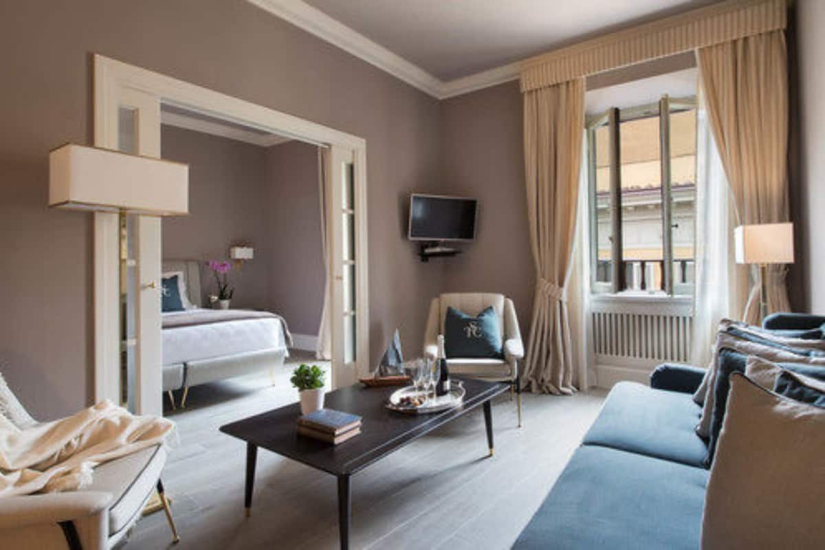 Quarto de hotel com cama, sofá, mesa de centro, televisão e janela com cortinas.