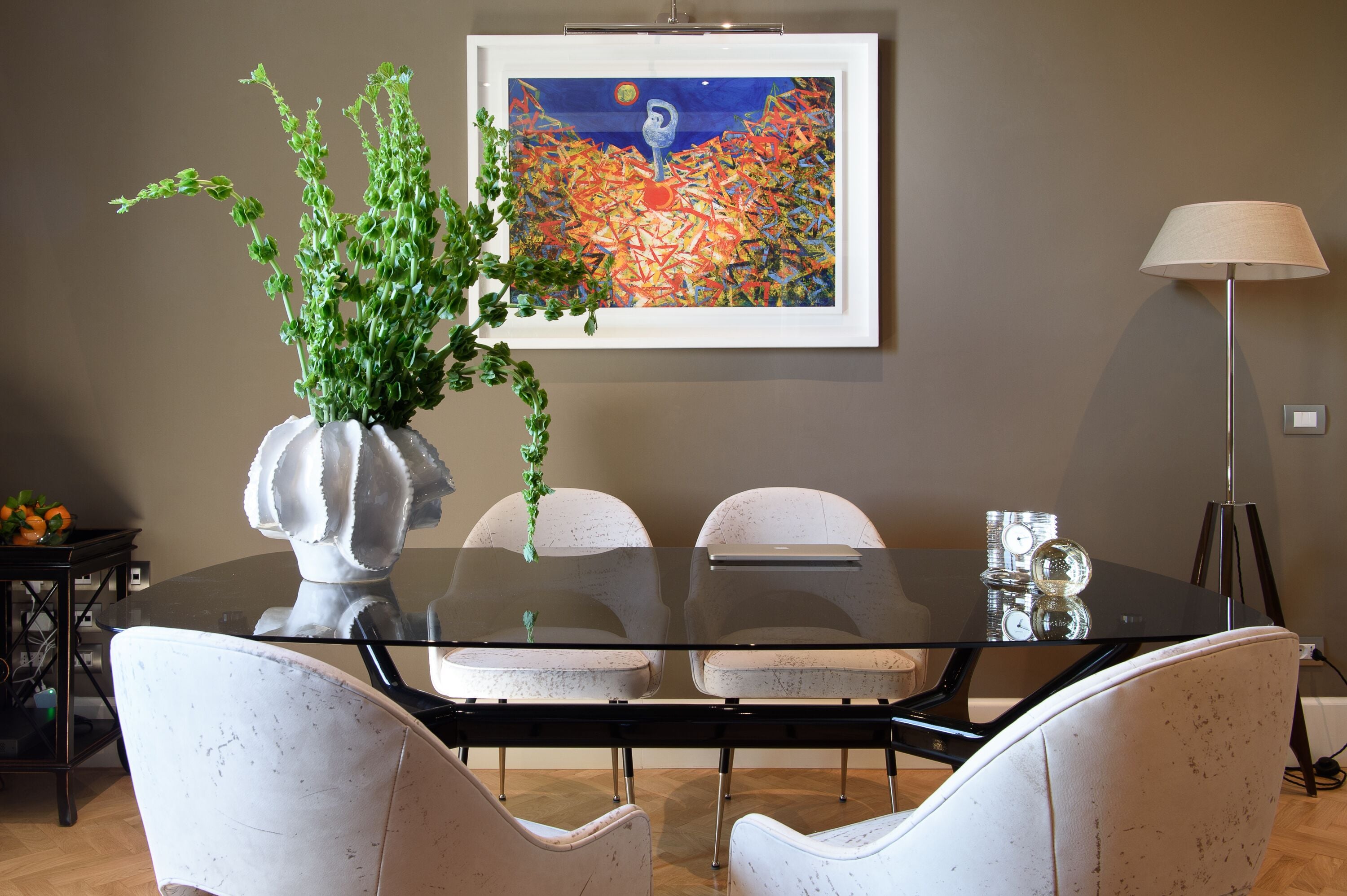 A dining area with a glass table, white chairs, a vase with green plants, a framed abstract painting, and a floor lamp.