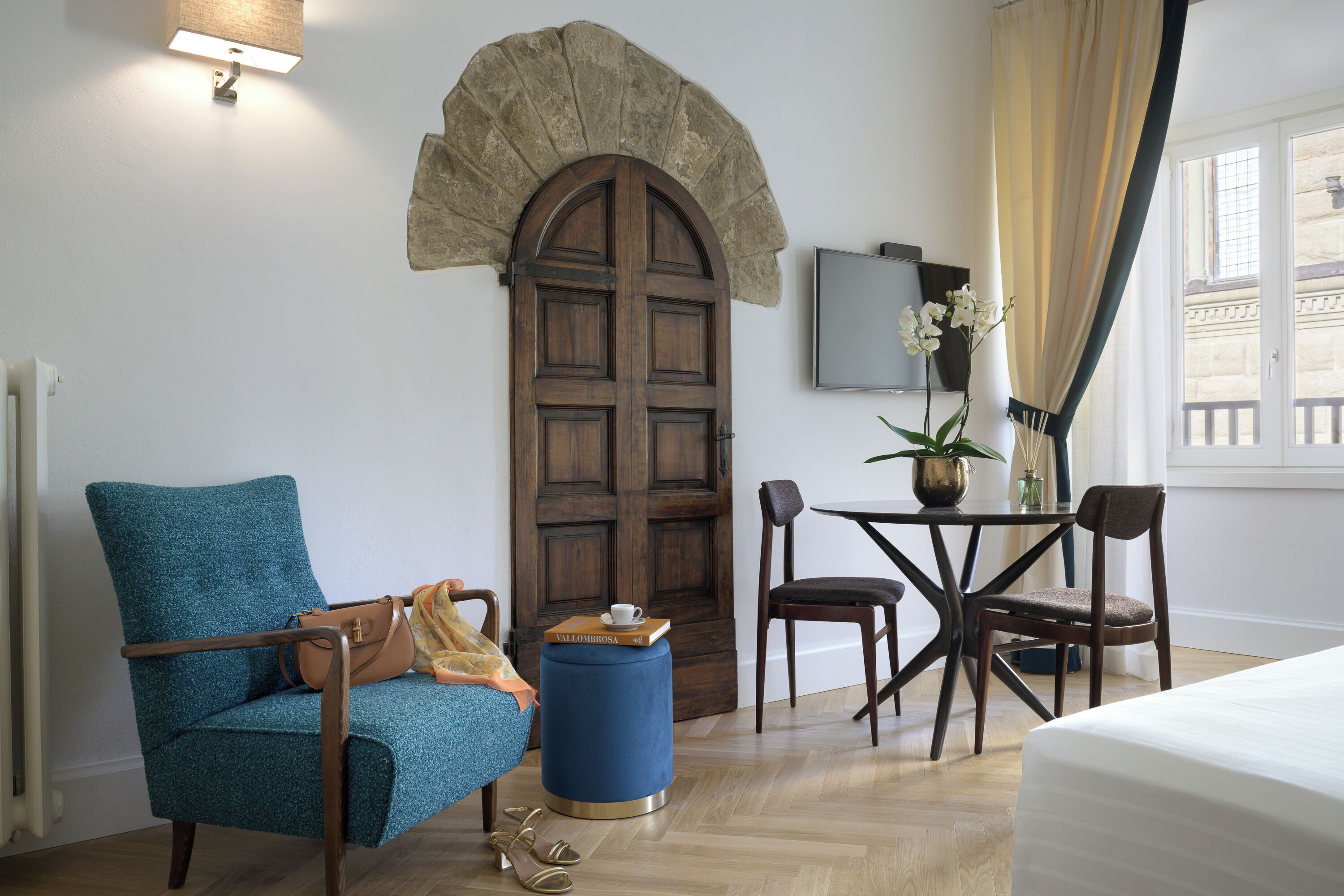 Una habitación de hotel con una puerta de madera grande, un sillón azul, una mesita redonda con un florero y una cama.