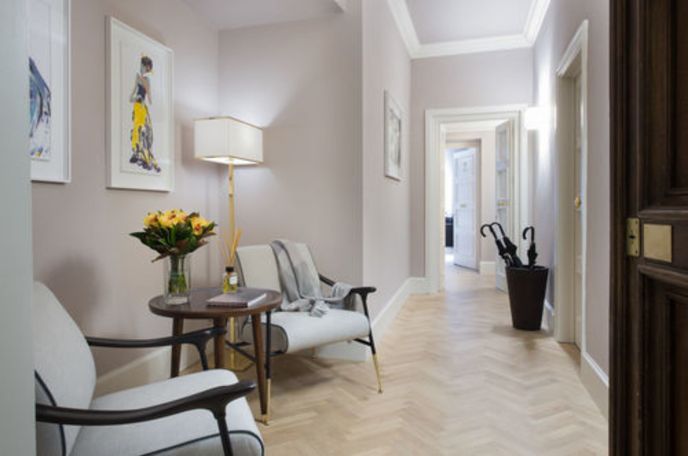 A hallway with a wooden floor, a table with a vase of flowers, two chairs, and framed artwork on the walls.