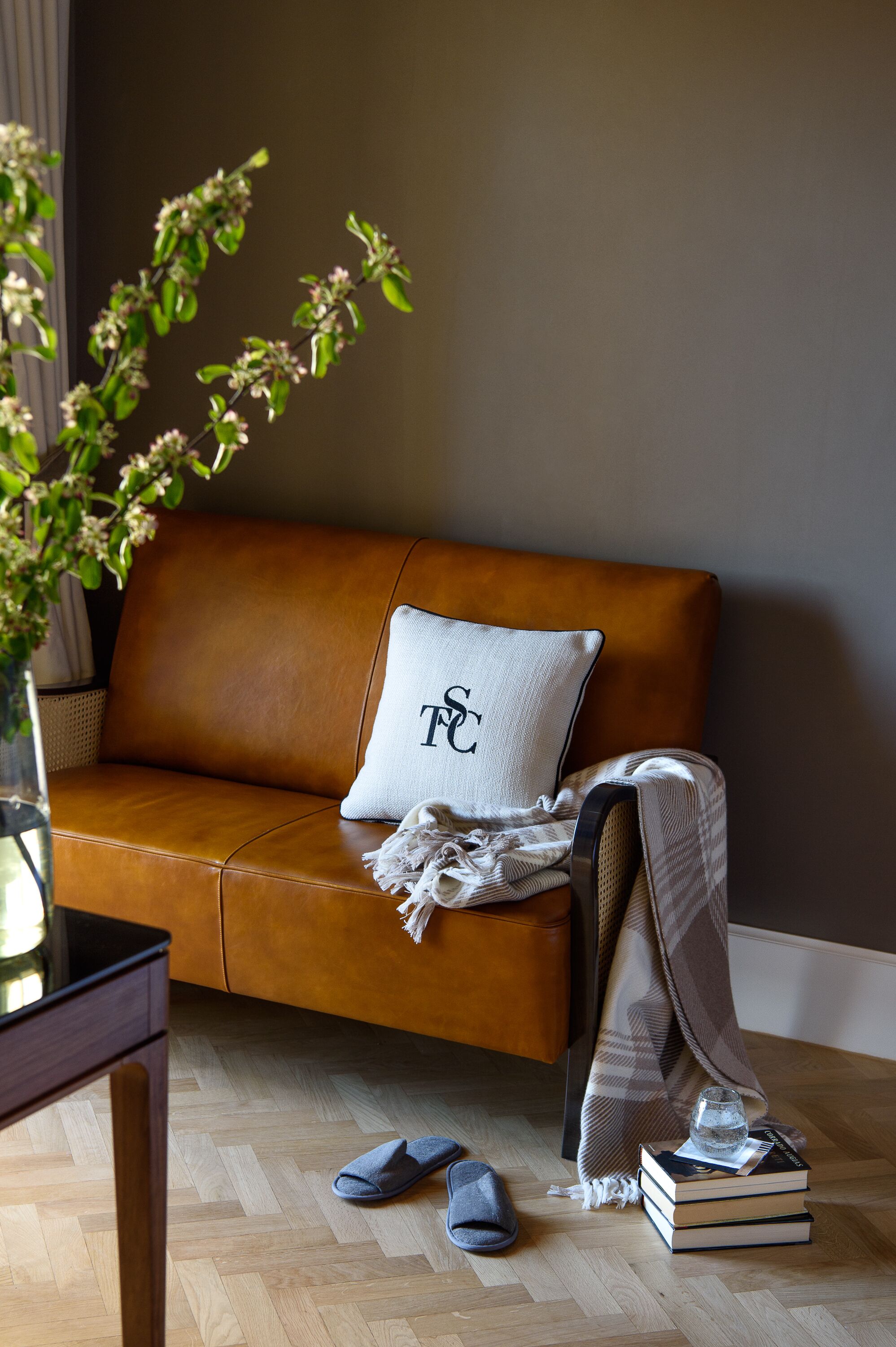 A cozy living room with a brown leather sofa, a white pillow with a logo, a blanket, a pair of slippers, and a stack of books.