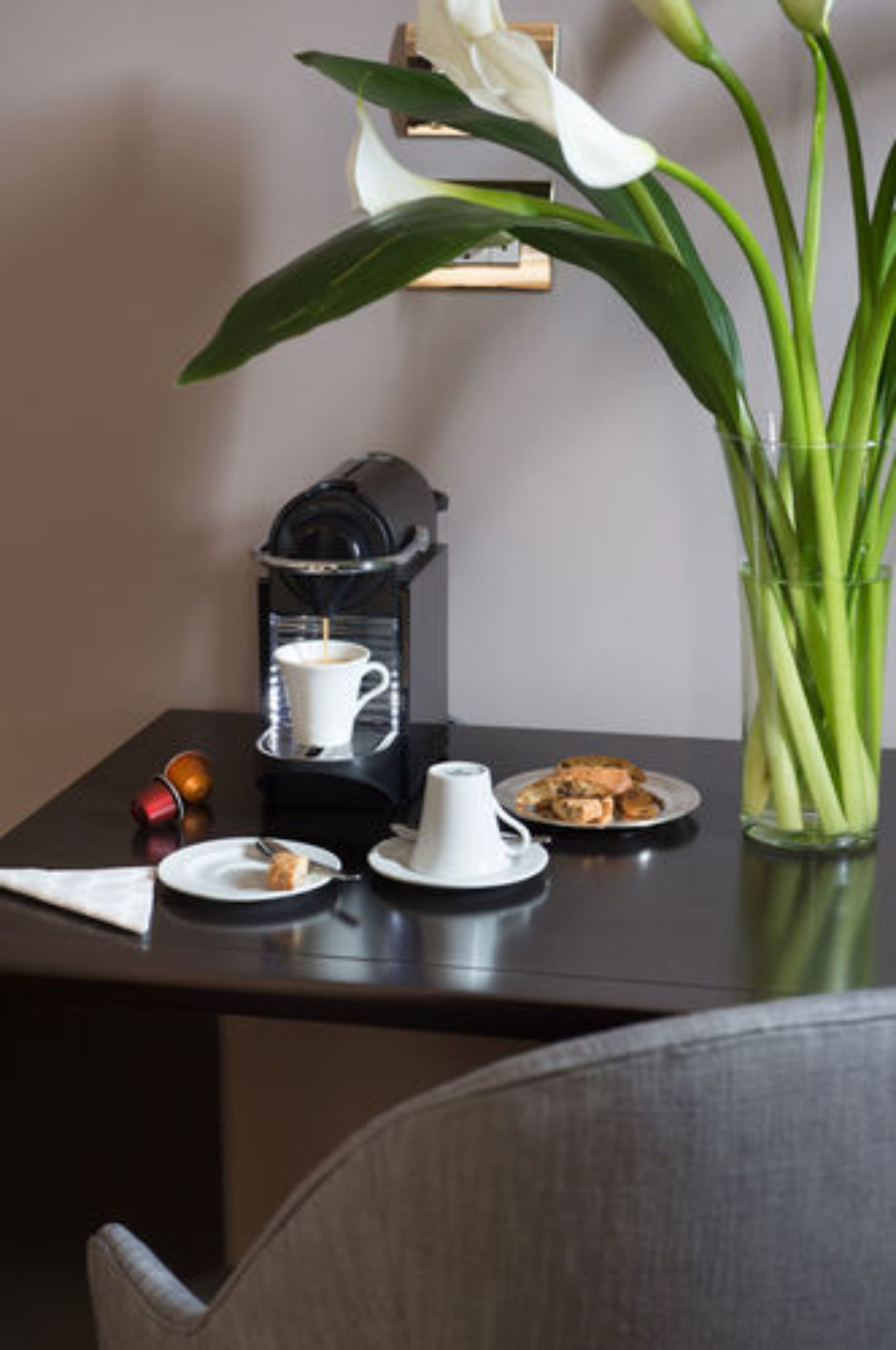 Une table avec une machine à café, une tasse de café, une assiette de biscuits, un vase de fleurs et une serviette.