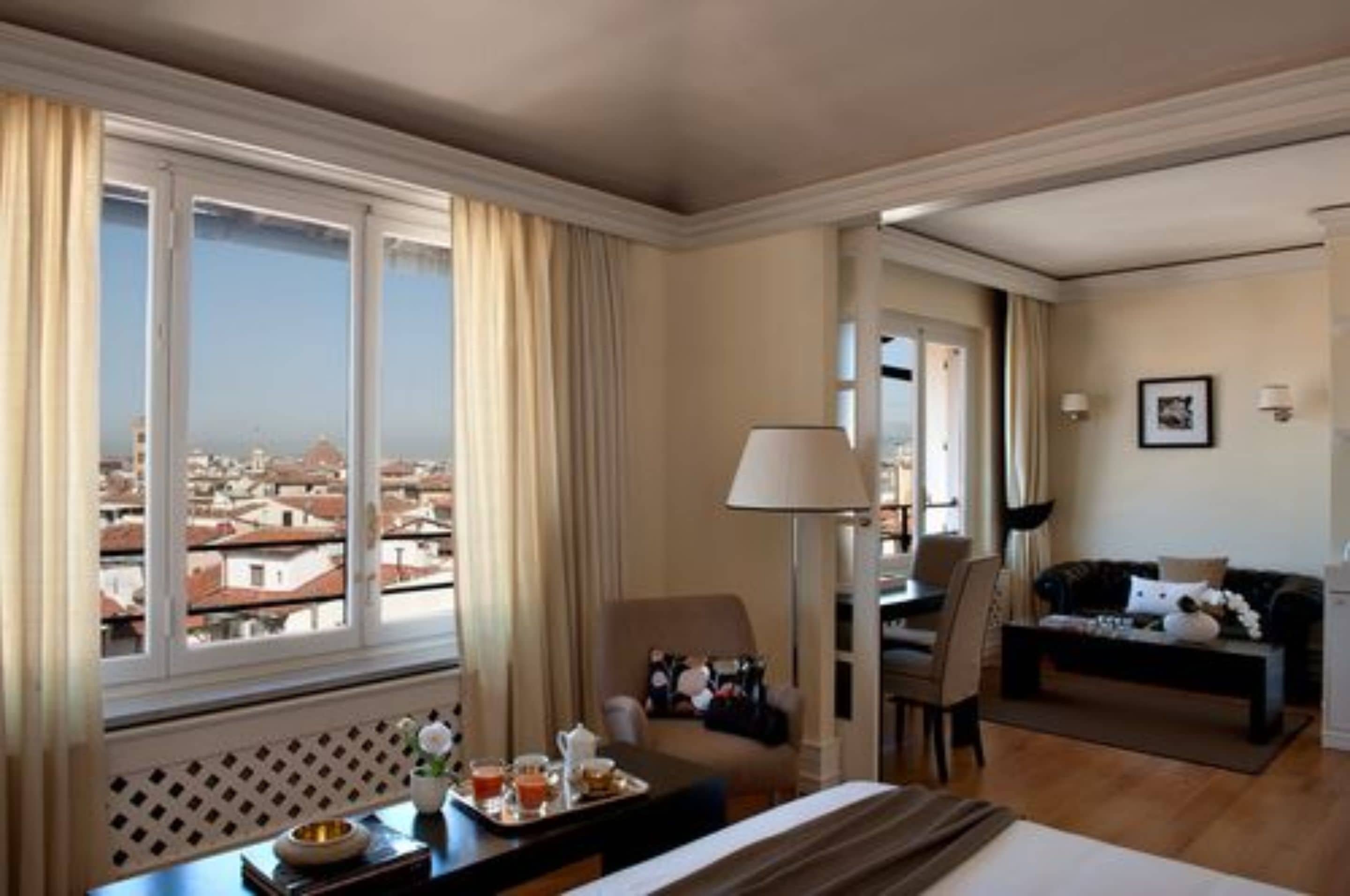 A hotel room with a large window, a sofa, a dining table, and a lamp.