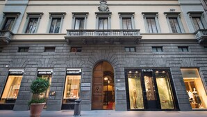 Un edificio de varios pisos con una entrada central, locales de Mont Blanc y Prada, y una planta grande en maceta al frente.
