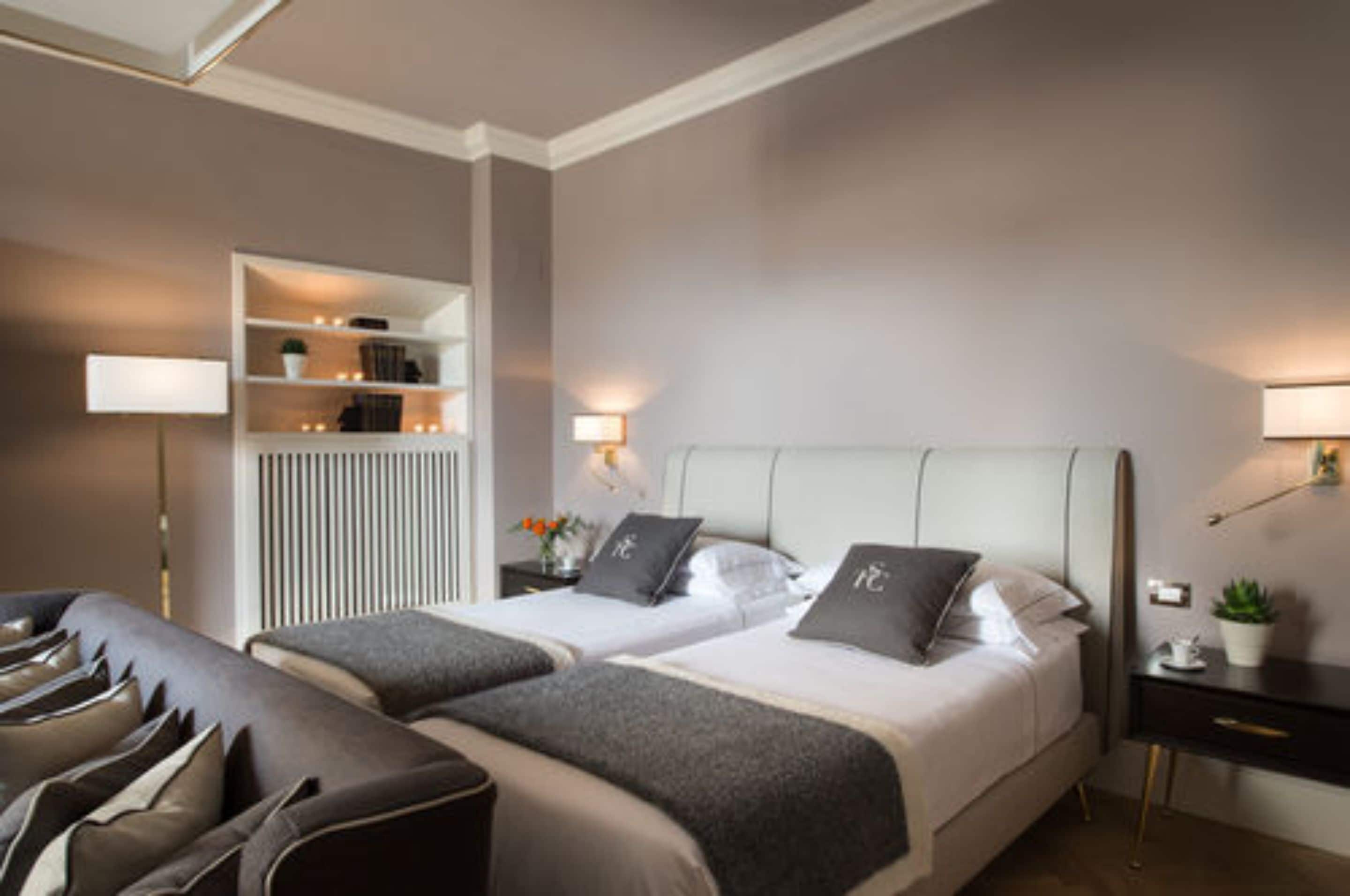 Moderni hotellihuone, jossa on kaksi sänkyä, oleskelualue, yöpöytä lampun kanssa ja pieni huonekasvi.