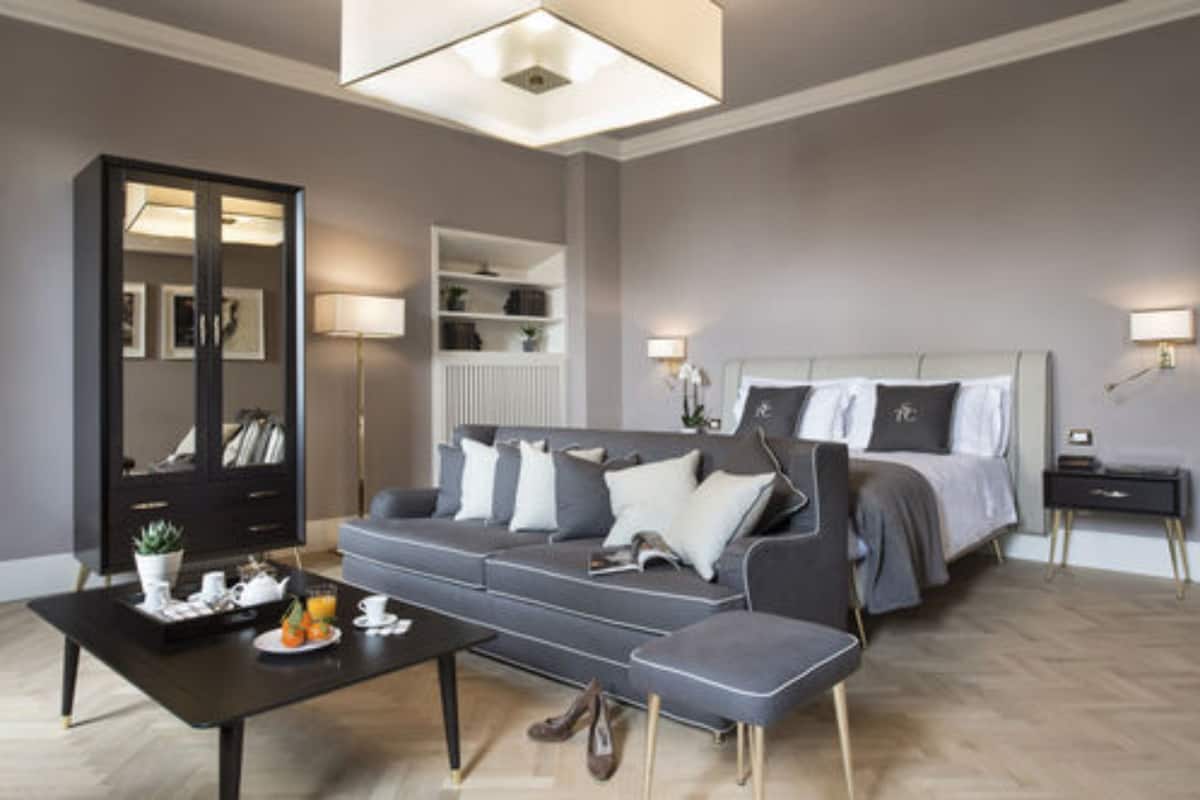 Quarto de hotel moderno com um sofá cinzento, uma cama com roupa de cama branca, uma mesa de cabeceira preta, um abajur e uma estante embutida.