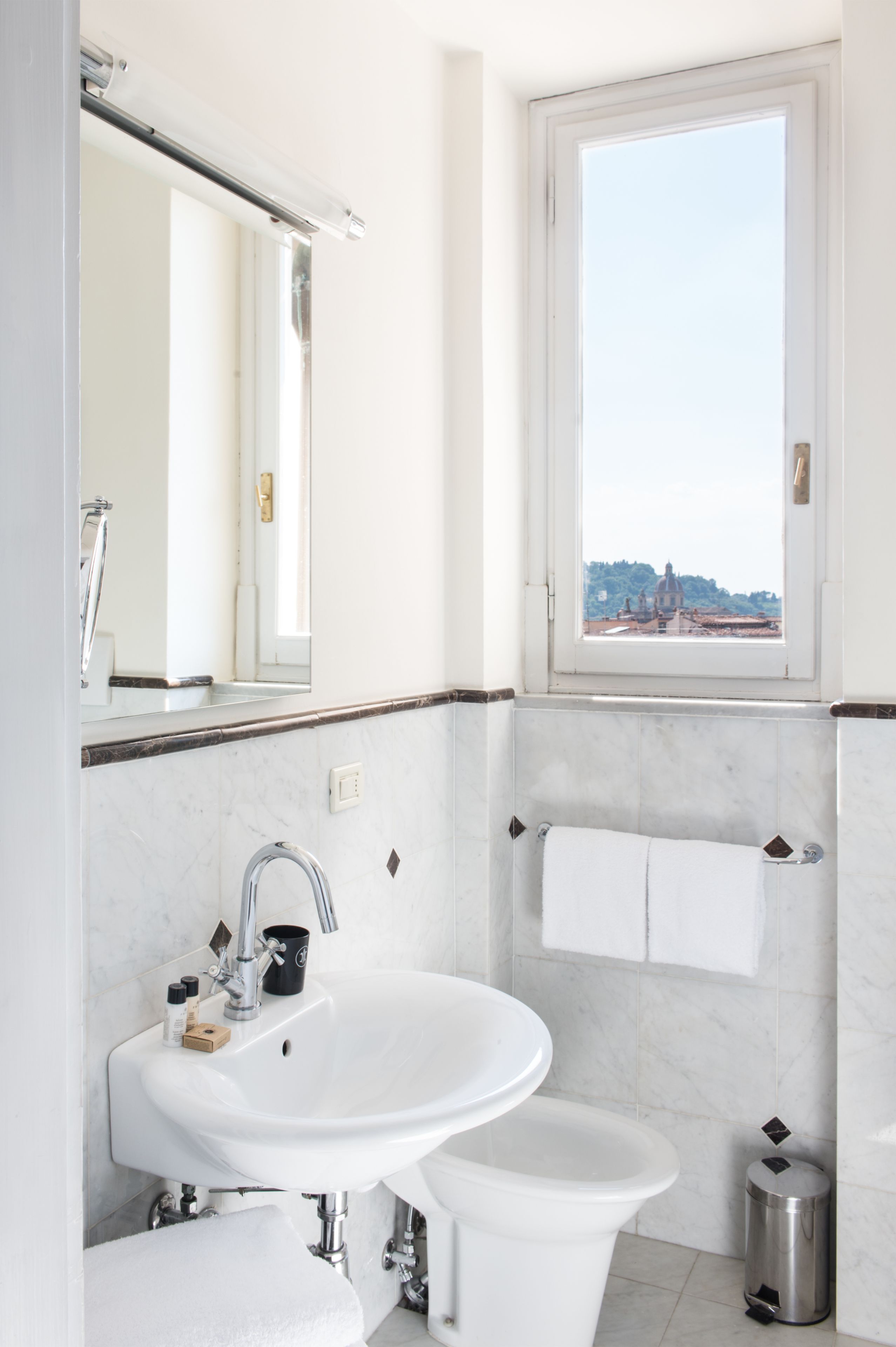 Un bagno con un lavandino bianco, uno specchio, una finestra e vista su un edificio.