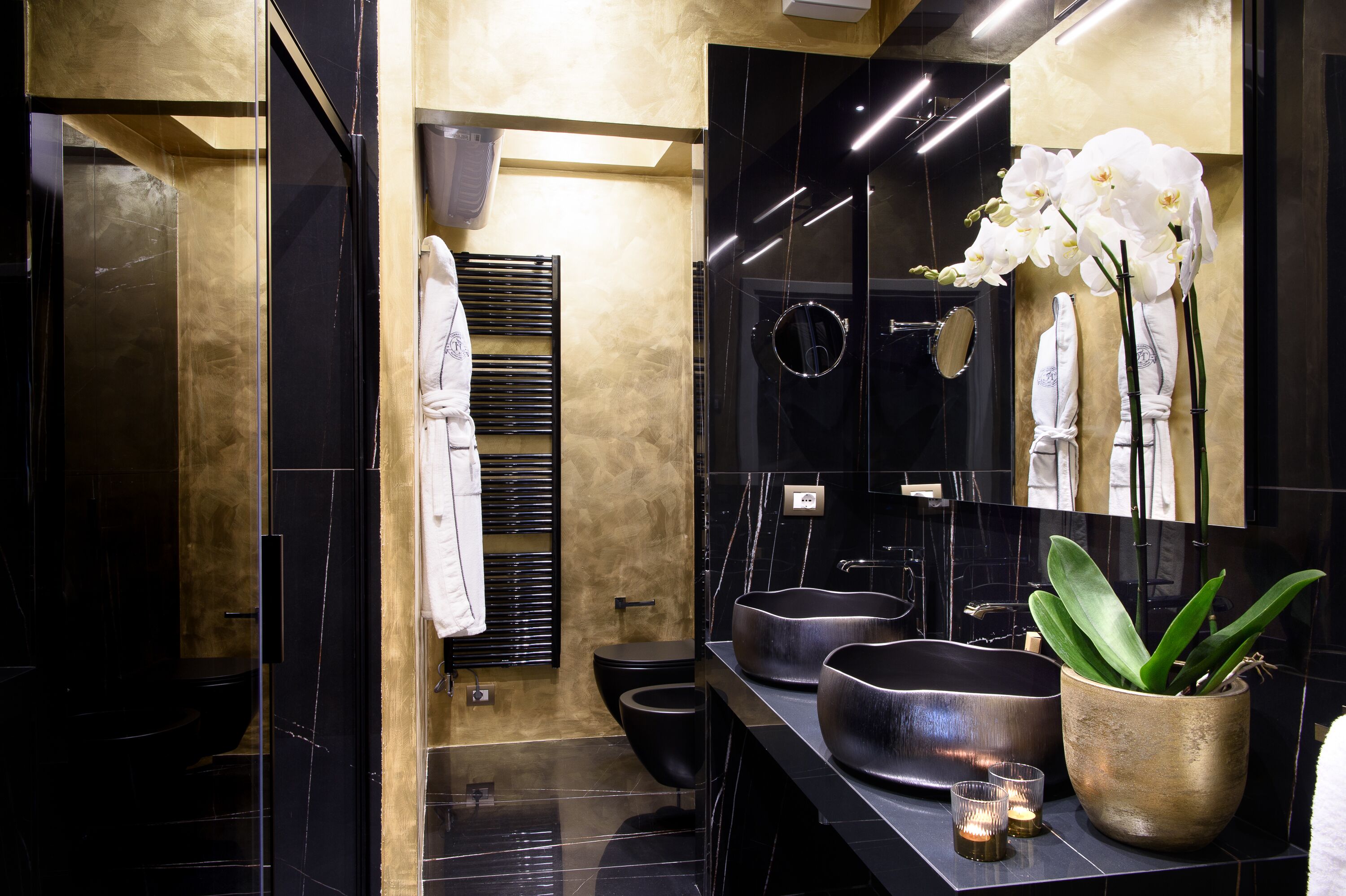 A modern bathroom with a black and gold color scheme, featuring a large mirror, two sinks, a toilet, and a potted plant.