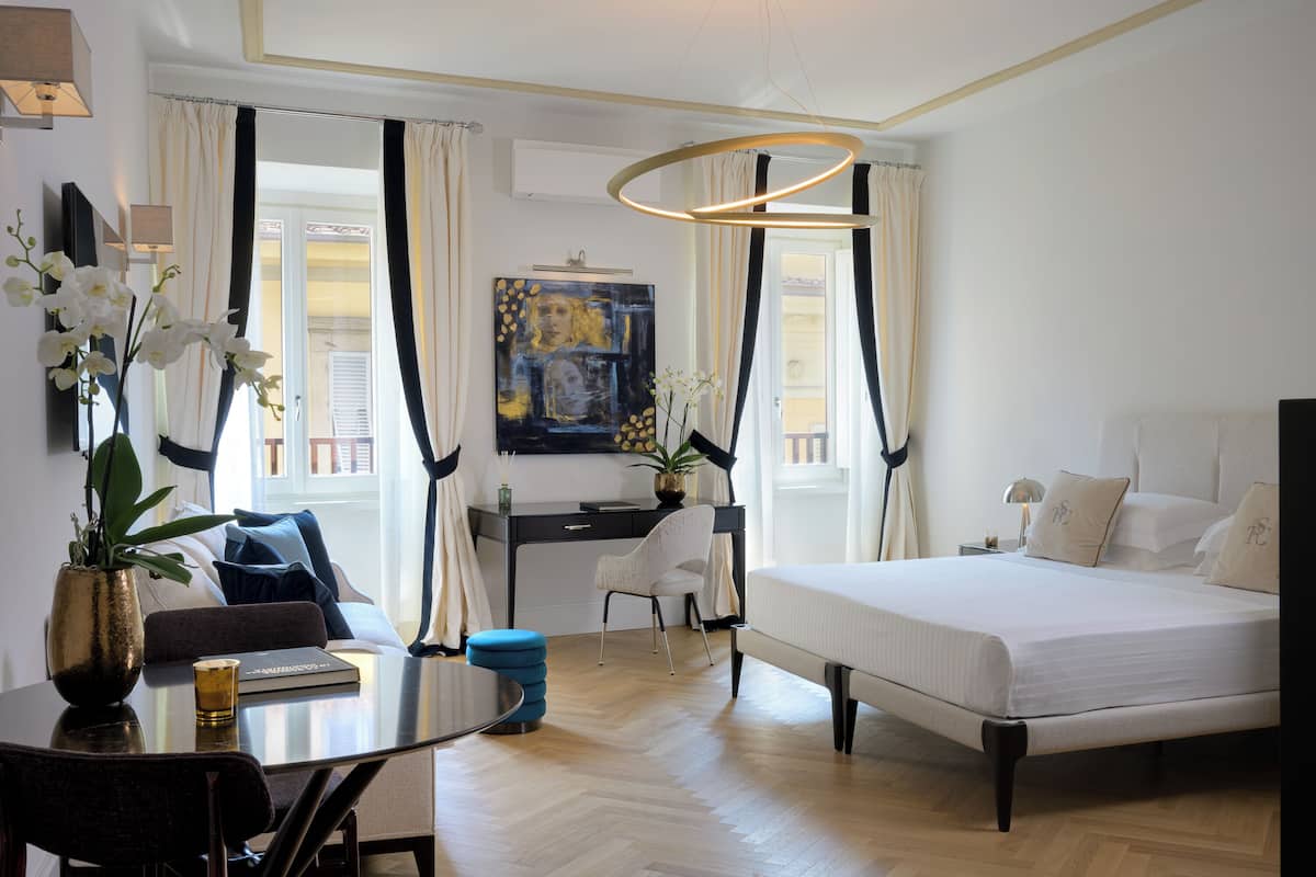 Quarto de hotel com cama, escrivaninha, cadeira, mesa com um vaso de flores e um quadro na parede.