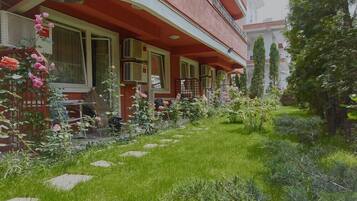 A building with a red facade, a covered outdoor seating area, and a garden with various plants and flowers.