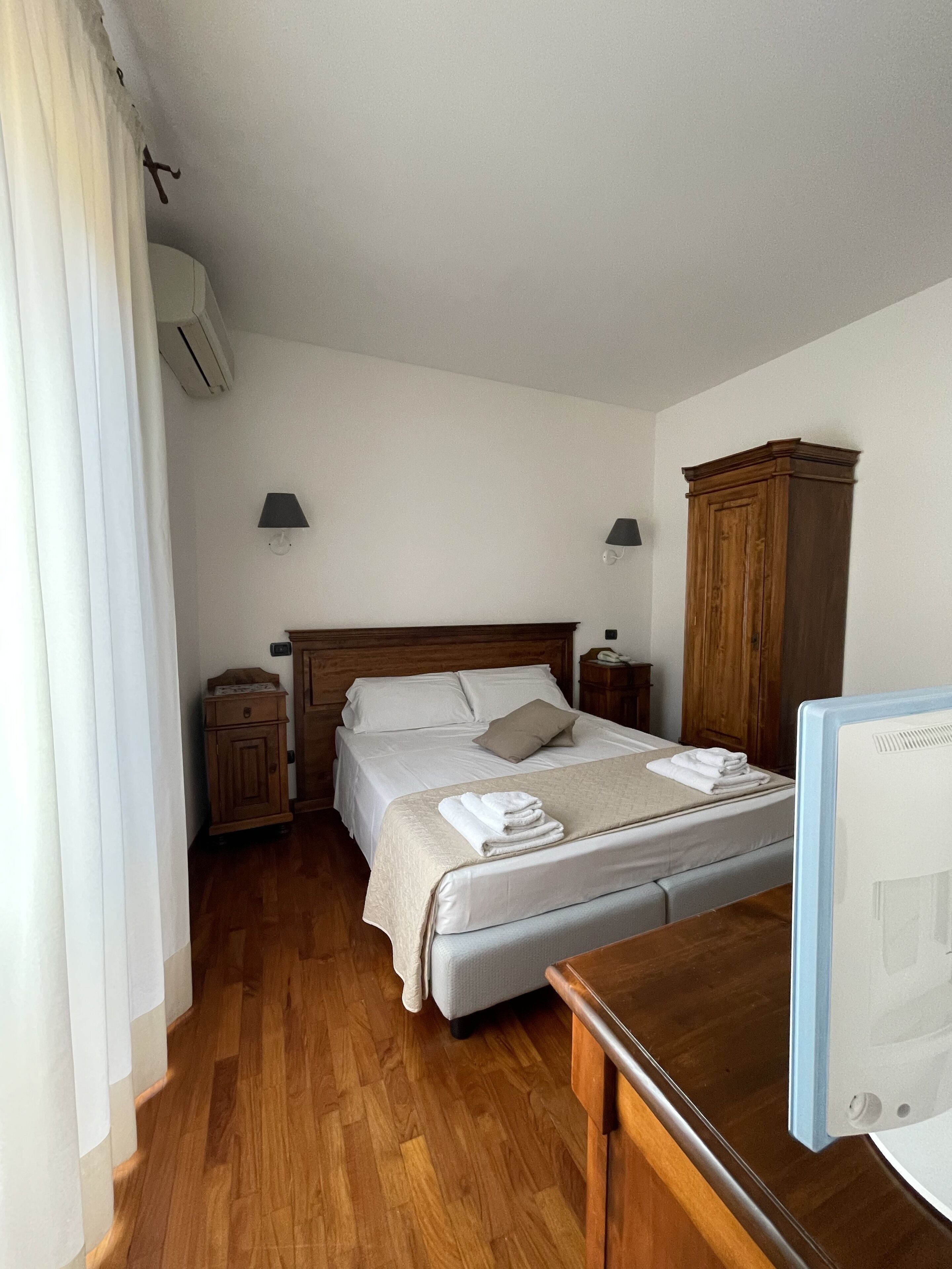 A hotel room with a wooden bed, a wooden wardrobe, a desk with a computer, and a window with curtains.