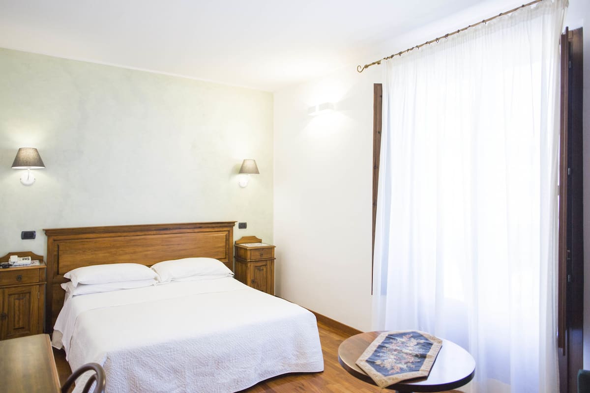 Ein Schlafzimmer mit einem hölzernen Bett, zwei Nachttischen, einem runden Tisch mit einem Buch und leicht transparenten Vorhängen.