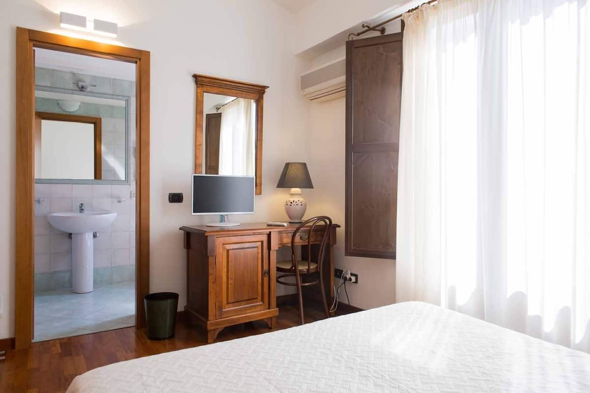 Ein Schlafzimmer mit Bett, Nachttisch mit Lampe, Stuhl, Schreibtisch mit Computer sowie ein Badezimmer mit Waschbecken und Spiegel.