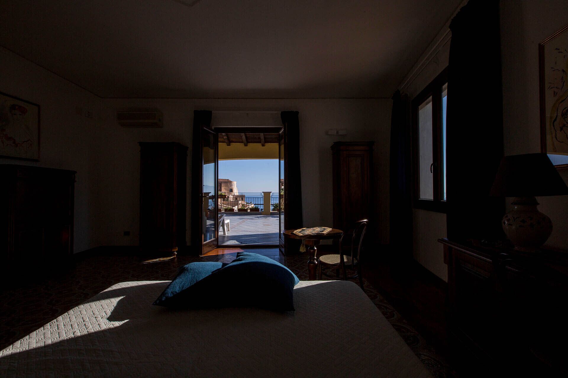 A dimly lit room with a bed, a small table, and a view of a courtyard and building outside.