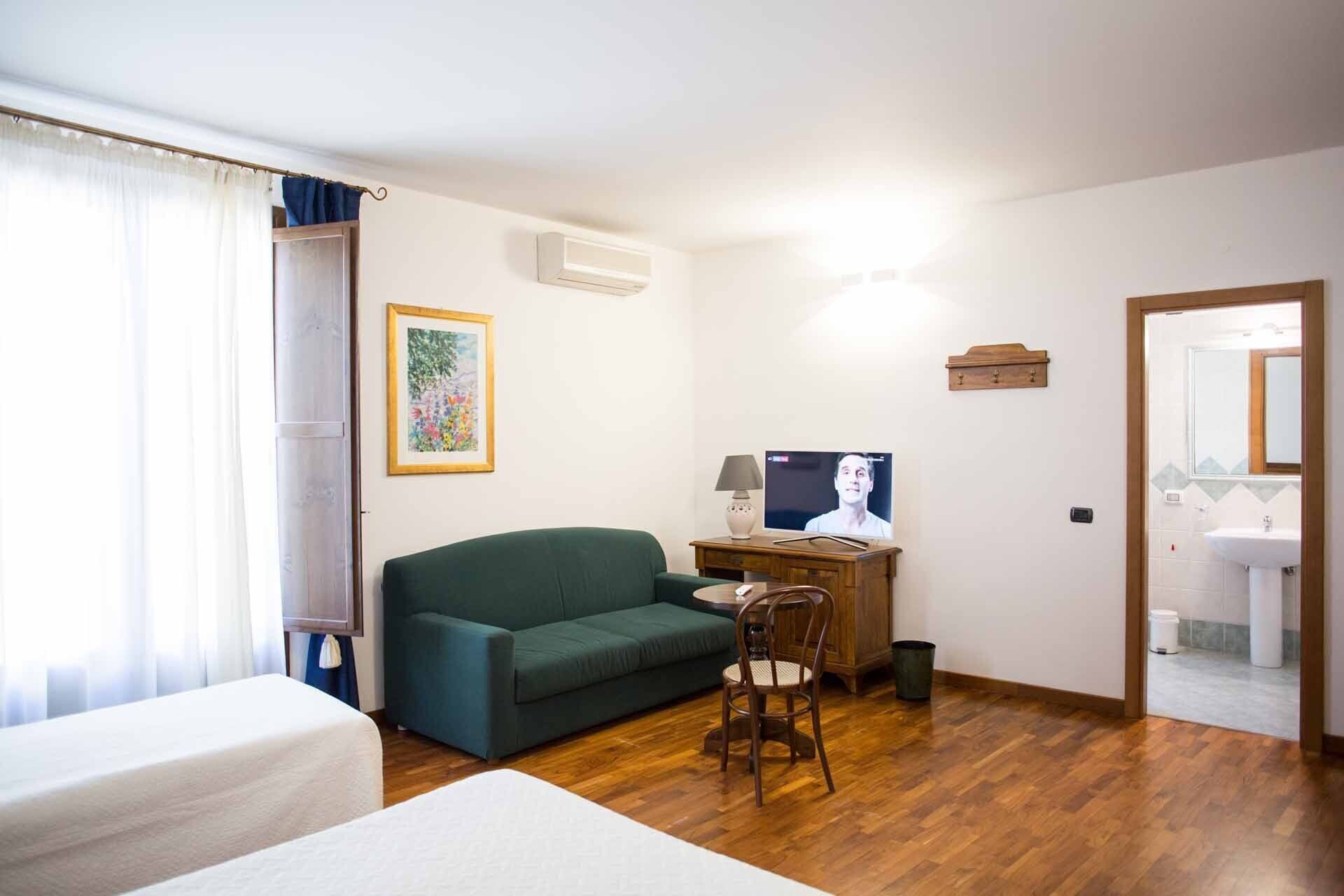 Een kamer met een groene bank, een klein houten tafeltje met een lamp, een lijst met een afbeelding aan de muur, en een badkamer zichtbaar door een geopende deur.