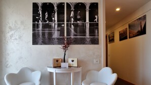 Un área de recepción con dos sillas blancas, una pequeña mesa con un florero y elementos enmarcados, y una fotografía grande en blanco y negro en la pared.