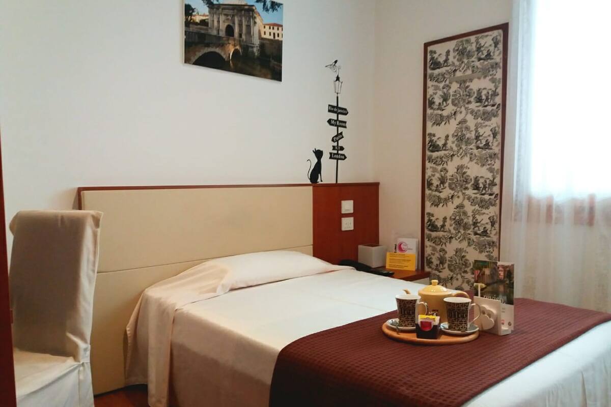 A hotel room with a bed, a chair, a tray with mugs, and a wall with a sign and a painting.