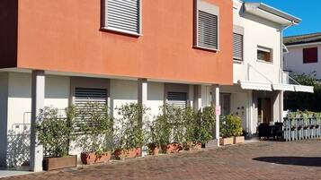 A building with a flat roof, two visible windows with shutters, and a paved area with potted plants.