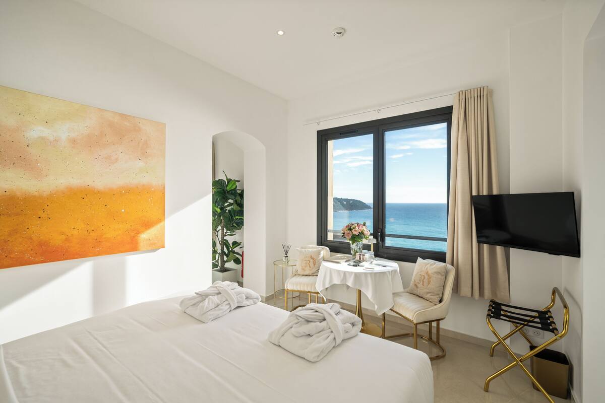 Ein Hotelzimmer mit einem großen Bett, einem Fernseher, einem Fenster mit Meerblick und einem Bild an der Wand.