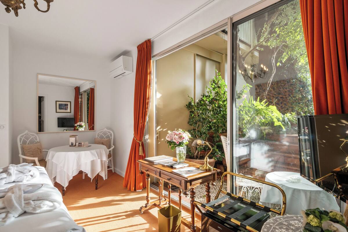 Ein Hotelzimmer mit einem Bett, einem Schreibtisch mit einem Laptop, einem Stuhl, einem Tisch mit Blumen und einem großen Fenster mit Vorhängen.