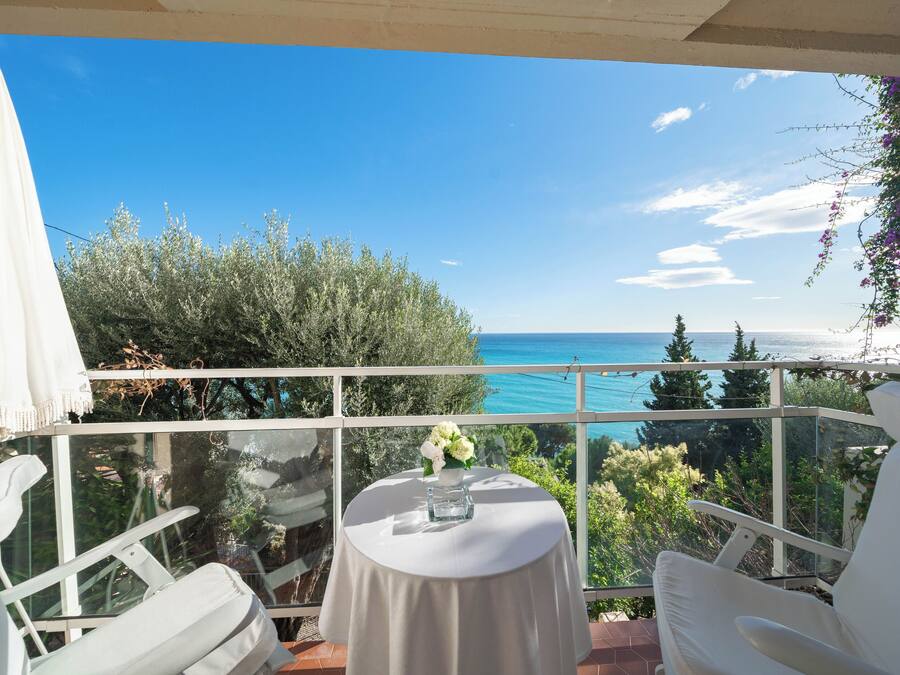 A balcony with a table set for two, overlooking a scenic view of the sea and greenery.