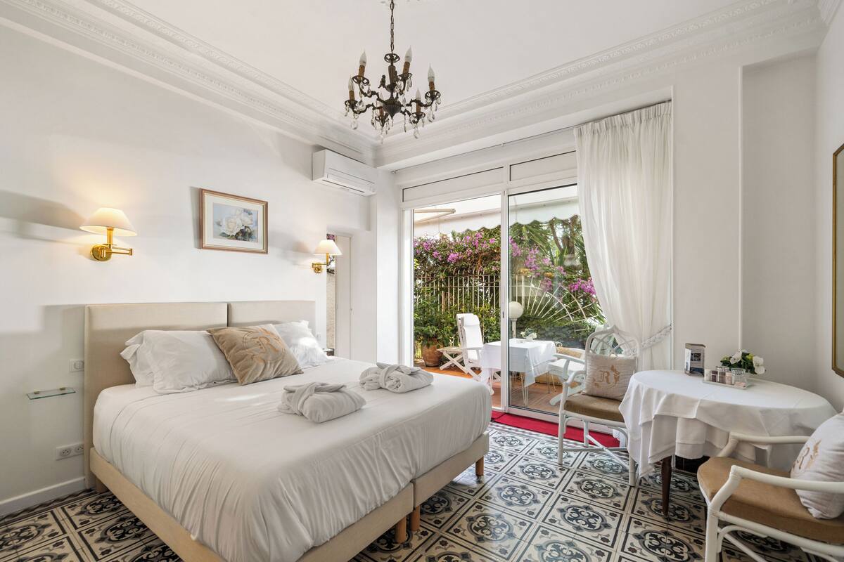 Ein Schlafzimmer mit einem Bett, Nachttischlampen, einer Deckenleuchte, einem Fenster mit Vorhängen, Blick auf den Garten und einem kleinen Tisch mit Stühlen.