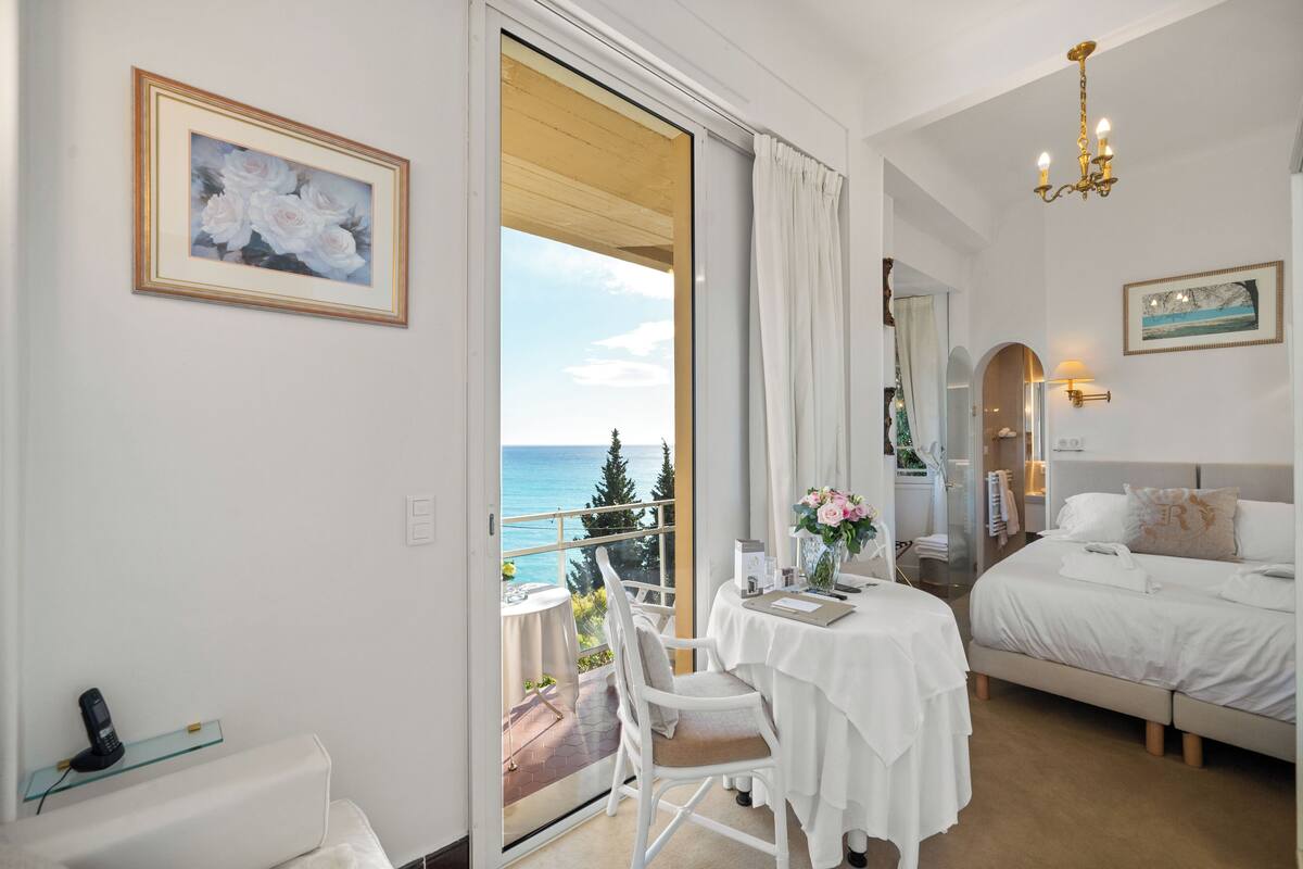 Ein Hotelzimmer mit Balkon, einem Bett, einem Tisch mit Blumenvase und einem Bild an der Wand.