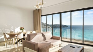 A modern living room with a large window overlooking the sea, a sofa, a dining table with chairs, and a coffee table. - Le Roquebrune (Roquebrune-Cap-Martin)