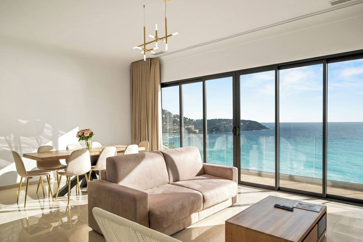 Ein modernes Wohnzimmer mit großem Fenster zum Meer, einer Couch, einem Esstisch mit Stühlen und einem Couchtisch.