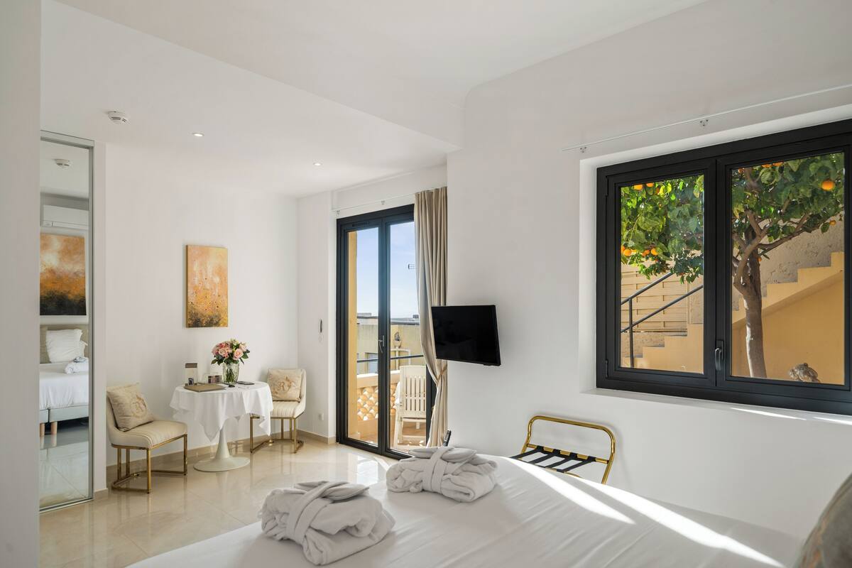 Ein Hotelzimmer mit Bett, Fernseher, Balkon mit Blick auf Bäume und ein Gebäude, Tisch mit Stühlen und einem Bild an der Wand.