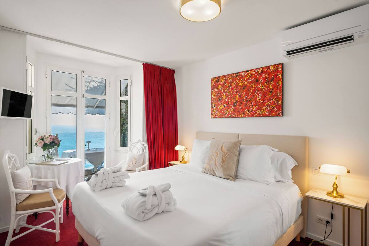 Ein Hotelzimmer mit einem großen Bett, zwei Nachttischen mit Lampen, einem Fernseher, einem Sessel, einem Tisch mit Blumen und Meerblick durch große Fenster.